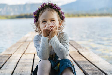 Nahaufnahme eines süßen Mädchens mit Diadem, das lachend auf einem Steg am See sitzt - DIGF12793