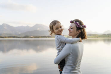 Smiling mother wearing tiara carrying daughter while standing against lake at sunset - DIGF12796