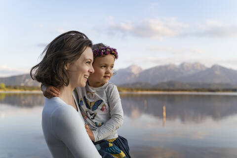 Smiling mother carrying cute daughter while standing against lake at sunset stock photo