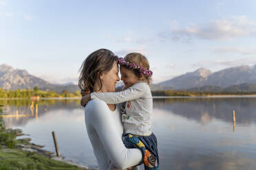 Mother carrying cheerful daughter while standing by lake against sky during sunset - DIGF12798