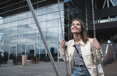 Smiling young woman sitting on swing against modern building in city - HPSF00039