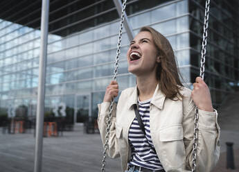 Happy young woman screaming while swinging against modern building in city - HPSF00040