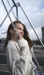 Happy young woman looking away while swinging against cloudy sky in city - HPSF00041