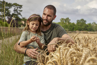 Father with daughter sitting by crops in wheat field against sky - VEGF02536