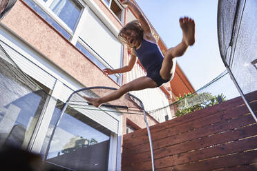 Fröhliches Mädchen springt an einem sonnigen Tag auf einem Trampolin vor einem Haus - VEGF02539