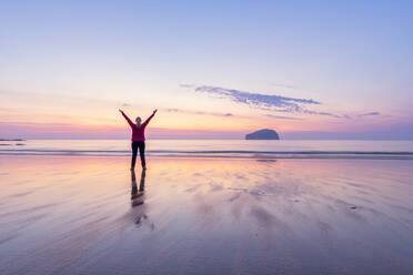 Mature woman with arms raised standing at Seacliff Beach during sunset, North Berwick, Scotland - SMAF01938