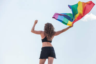 Mittlere erwachsene Frau winkt mit Regenbogenflagge und steht gegen den Himmel - MRRF00150