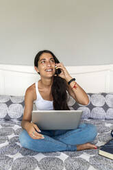 Lächelnde Frau im mittleren Erwachsenenalter, die über ein Smartphone spricht, während sie mit einem Laptop auf dem Bett sitzt - EGAF00531
