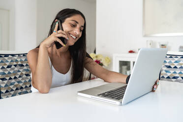 Lächelnde Frau im mittleren Erwachsenenalter, die über ein Mobiltelefon spricht, während sie einen Laptop auf einem Tisch zu Hause benutzt - EGAF00542