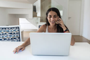 Lächelnde Frau, die über ein Mobiltelefon spricht, während sie einen Laptop auf dem Tisch benutzt - EGAF00544