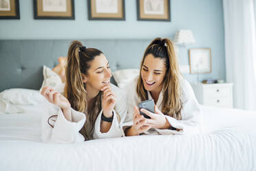 Lächelnde Zwillingsschwestern, die ein Smartphone benutzen, während sie auf dem Bett im Hotel liegen - OCMF01557
