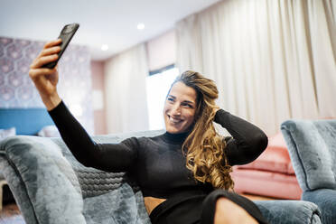 Lächelnde Frau, die ein Selfie mit ihrem Smartphone macht, während sie auf einem Sessel im Hotelzimmer sitzt - OCMF01566