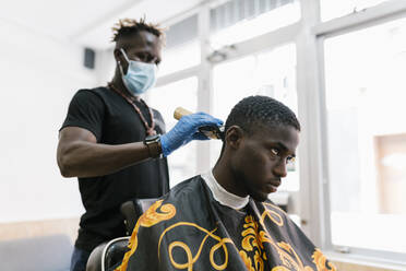 Friseur mit Handschuhen und Maske schneidet die Haare eines jungen Mannes mit einem Rasiermesser in einem Salon - EGAF00562