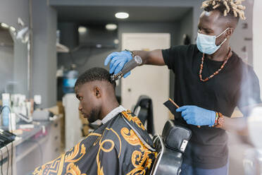Friseur mit Locken, der Maske und Handschuhe trägt, während er die Haare eines Kunden im Salon schneidet - EGAF00565