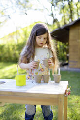 Nettes Mädchen pflanzt Samen in kleine Töpfe, während sie an einem Tisch im Garten steht - BRF01477