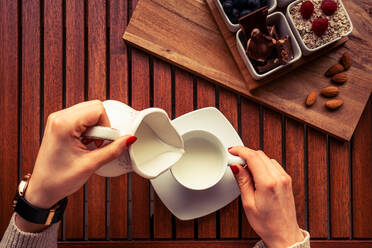 Hands of anonymous woman filling cup with fresh dairy near set of assorted food for breakfast on timber tabletop - ADSF04376