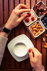 Hands of anonymous lady holding cup of fresh milk and taking ripe raspberry from bowls with assorted breakfast food over wooden table - ADSF04377