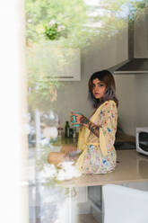Hübsche junge Frau im Seidengewand, die eine Tasse mit einem frischen Heißgetränk hält und in die Kamera schaut, während sie auf dem Küchentisch hinter dem Fensterglas sitzt - ADSF04399
