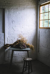 Konzept eines Blumenstraußes aus trockenen Nadelbaumzweigen in Bastelpapier auf einem Tisch in der Nähe eines Hockers in einem grauen, trüben Raum mit Backsteinwänden - ADSF04439