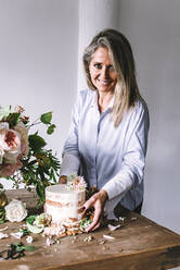 Seitenansicht der Dame Platzierung Gericht mit leckeren Kuchen dekoriert Blüte Knospe auf Holztisch mit Strauß Chrysanthemen, Rosen und Pflanze Zweige in Vase zwischen trockenen Blättern auf grauem Hintergrund - ADSF04442