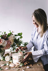 Seitenansicht der Dame Platzierung Gericht mit leckeren Kuchen dekoriert Blüte Knospe auf Holztisch mit Strauß Chrysanthemen, Rosen und Pflanze Zweige in Vase zwischen trockenen Blättern auf grauem Hintergrund - ADSF04443