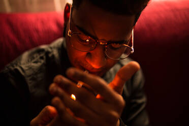 Close-up of young trendy black man in eyeglasses lighting cigarette while sitting on red - ADSF04521