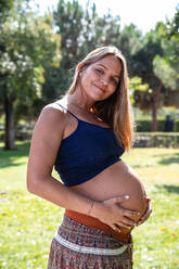Seitenansicht junge fröhliche schwangere Frau in Rock und Tank Top hält Bauch in grünen Garten - ADSF04550