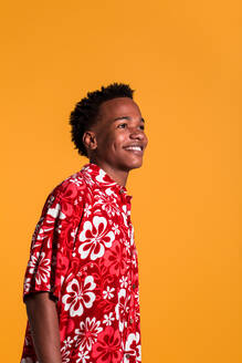 Seitenansicht eines lächelnden jungen afroamerikanischen Mannes, der ein buntes Strandhemd trägt und auf einem orangefarbenen Hintergrund wegschaut - ADSF04574