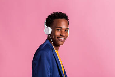 Seitenansicht eines jungen schwarzen Mannes in bunter lila Jacke und?mit Kopfhörern, der in die Kamera lächelt, auf rosa Hintergrund - ADSF04581