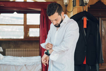 Young handsome man with dark hair and beard putting on white shirt in bedroom with new black costume hanging on clothes hanger - ADSF04612