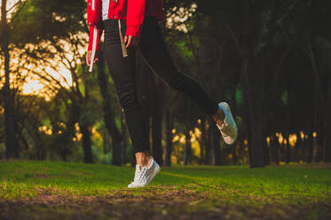 Crop shot of woman in sportive jacket and sneakers jumping slightly standing on tiptoe on green grass in park - ADSF04623