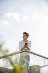 Handsome young guy in casual outfit browsing smartphone and looking away while leaning on railing on background of cloudy sky - ADSF04633