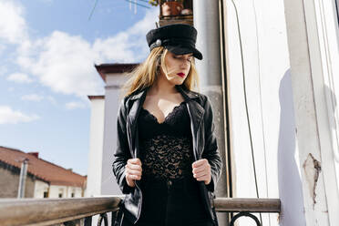 Side view of attractive young female in stylish hat and leather jacket while standing on balcony railing on sunny day in city - ADSF04650