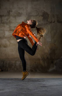 Side view of young lady in casual wear jumping with leg stretched to back in grey gloomy room - ADSF04653