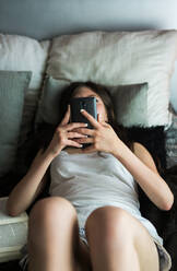 Teenager-Mädchen in Schlafanzug schaut nachmittags in ihrem Zimmer auf ihr Smartphone - ADSF04698