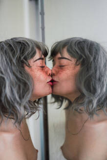 Seitenansicht einer jungen Frau mit nackten Brüsten, die die Augen geschlossen hält und das Spiegelbild im Spiegel küsst - ADSF04772