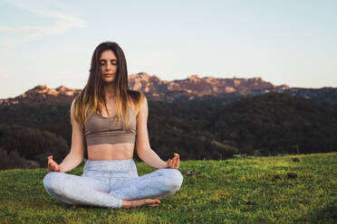 Junge Frau in Sportkleidung sitzt auf einer grünen Wiese und macht mit geschlossenen Augen Yoga. - ADSF04788