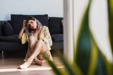 Inked woman adjusting hair and looking at camera while sitting on floor at couch - ADSF04800