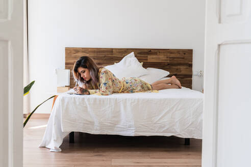 Seitenansicht einer niedlichen jungen Frau im Seidenmantel, die auf einem bequemen Bett liegt und in einem kleinen Notizblock Skizzen anfertigt, während sie ihre Zeit in einem stilvollen Schlafzimmer verbringt - ADSF04807