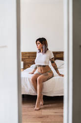 Attractive tattooed lady in panties and T-shirt sitting on comfortable bed and looking away - ADSF04809