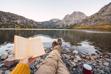 Crop Beine des Menschen hält Volumen in der Nähe Becher auf Stein Küste in der Nähe von Wasser von Mammoth Lakes und malerischen Blick auf Berge - ADSF04816