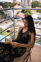 Schöne leidenschaftliche brunette Frau im Kleid sitzt auf Stuhl mit Volumen auf Balkon in der Nähe von modernen Gebäuden - ADSF04823