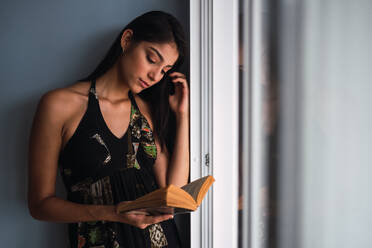 Schöne leidenschaftliche brünette Hispanic Frau im Kleid Blick auf Band in den Händen in der Nähe von Fenster und graue Wand - ADSF04824