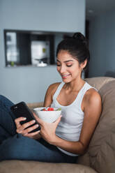 Beautiful brunette Hispanic woman holding fork with slice of fruit and sitting on settee with bowl of kiwi and watermelon and browsing in mobile phone - ADSF04832
