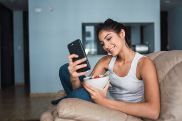 Beautiful brunette Hispanic woman holding fork with slice of fruit and sitting on settee with bowl of kiwi and watermelon and browsing in mobile phone - ADSF04833