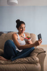 Beautiful brunette Hispanic woman holding fork with slice of fruit and sitting on settee with bowl of kiwi and watermelon and browsing in mobile phone - ADSF04834