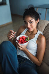Schöne brünette hispanische Frau, die eine Gabel und eine Schale mit Kiwi und Wassermelone hält und auf einem Sofa sitzt - ADSF04835