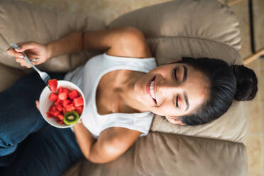 Beautiful cheerful brunette Hispanic woman holding fork and bowl of kiwi and watermelon and sitting on settee - ADSF04836