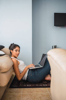 Schöne brünette hispanische Frau, die auf einem Laptop tippt und auf dem Boden auf einem kleinen Teppich in der Nähe des Sofas und des Fensters sitzt - ADSF04838