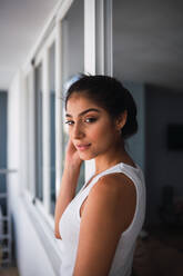 Beautiful brunette thoughtful Hispanic woman in casual wear standing between balcony windows - ADSF04839